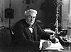 Black and white photograph of a white haired man in a suit and bow tie at a desk with several pieces of paper, right quarter profile, looking towards the viewer, holding a pen in the right hand