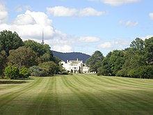 A white, two-storey building photographed with trees in the foreground and a mountain topped by a tower in the background.