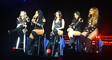 Five young women wearing black clothing sitting on tall chairs in a line on a stage.