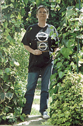 A middle-aged woman stands on a path amidst green vegetation, under bright sunlight. She wears a black T-shirt with a white logo, and holds a small piece of black card which carries the same logo. She has blue jeans, and white shoes. Her hair is cut short. She is looking slightly upward, over the head of the photographer.