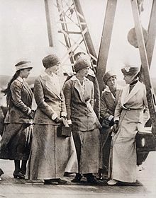 Three young ladies in the centre of the picture wear ankle-length skirts, matching jackets, and brimless hats, and look at an older lady on the right who wears similar clothes but with a veil. Their hair is pinned up beneath the hats. On the left a girl with loose hair, which streams down her back to just below her elbow, also looks towards the older lady. The girl wears a similar jacket, but with a skirt cut to just below the knee.