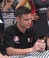 A man in his early twenties looking to someone outside the camera. He is wearing a black T-shirt with sponsors logos and his holding both his hands together/