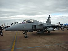 Three quarter view of grey jet fighter fitted with external fuel tanks on dirt-colored ground