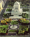 Aerial view of square white limestone building with pyramidal roof on a plaza; before it is a black obelisk with a golden capstone at the center of a circle