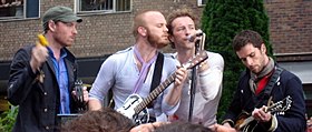 A color photograph of four members of Coldplay performing with instruments in front of a brick building