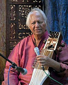 An old man sitting in front of an ornamented partition wall speaks into a microphone and holds a bowed instrument.