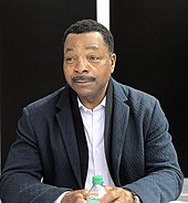 A man with black hair and a black moustache, wearing a black blazer and white patterned shirt, holds a water bottle and looks at something off-camera.