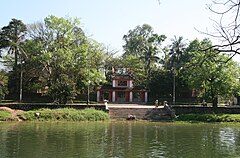 A green colored pond is at the front of a temple entrance. From the pond, there are paved stone steps up to the front of the temple, which is surrounded by a stone wall and a red triple gate with two levels and tiles. The area is surrounded by green shrubs and trees.