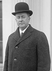 Cleanshaven man with drooping eyelids, at about 40. He is wearing a black bowler hat, white shirt, tie and dark overcoat