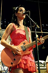 A brunette female in a red dress strums a guitar and sings into a microphone while performing on stage.