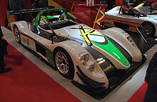 A green, black and white Formula 1 style race car from the front left, indoors on a red and gray carpet with other race cars in the background.