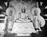 Front view of a central figure sitting cross-legged on a raised platform which is flanked by two smaller standing statues. The central figure has the palm of his right hand turned to the front. The attendants look identical pointing upwards with their right hand and their left hand lifted halfway touching the thumb with the middle finger. Each of the three statues has a halo. Black and white picture.