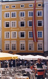 Facade of a tall brick building with rows of windows extending to five floors. Above the second row a sign indicates that this is Mozart's birthplace. In the foreground are sunshades and tables belonging to the modern café.