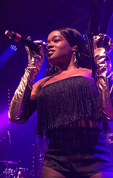 A woman on-stage, wearing metallic-coloured gloves that extend to her elbows, holds a mic in one hand.