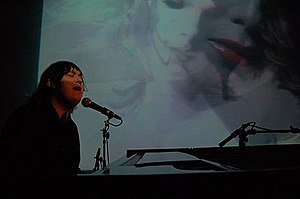 Antony Hegarty in 2008, at a grand piano singing into a microphone, in a black suit with hes hair lank, against a projected-visual backdrop.