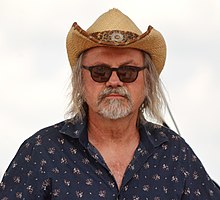 Ondřej Hejma wearing a dark blue patterned shirt, straw cowboy hat, and black sunglasses, looking directly at camera