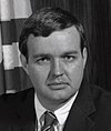 Black-and-white photo of a man in a suit and combed-over hair with the US flag behind him