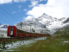 Red train with glass-canopied cars heading toward snow-capped mountains