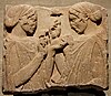 A marble fragment depicting two women holding their hands upraised with what appear to be flowers and other objects