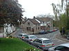 Street scene showing road junction and grey stone buildings with parked cars in front of them. To the left is a grassy area with a tree.