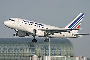 Mostly white aircraft with gears extended on takeoff. In the background are buildings.