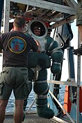 A diver in an armoured diving suit stands on a launch and recovery platform on the support vessel, attended by a crewman.