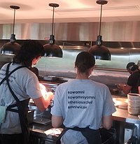 Two waitpersons and one chef face away from camera, bowls on stainless steel counter, three pendant lights hang from ceiling