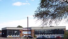 A building approximately 3 stories high. For the most part, the building is made up of horizontal columns of an opaque white panel up to around head height, with 3 windows arranged vertically above this. The columns are approximately 0.5m (1.5ft) wide and run from just below the roof to just above ground level and are separated by vertical wooden beams. Above the columns of windows a corrugated white border runs along the top of the building.