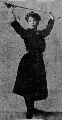 A white woman wearing a dark dress, holding a fencing pose