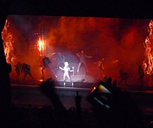 Faraway image of a stage lit by red light and backdrops of flames. A potlight falls on a female blond woman in the centre who wears a large white headdress and holds a stick in her left hand. She is surrounded by similar dressed male dancers who also wear a half-sleeved jacket.