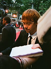 A picture of a man with red hair looking downward. A person is extending their arm to whist holding sheets of paper. Trees and red heights can be seen in the background.