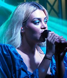 A 21-year-old woman is shown in an upper shot. She is holding a microphone on its stand with her right hand while her left is at the other end on the mike. She has her head turned to her left and is looking over her shoulder. She wears a white top and her brown hair is shoulder length. Behind her is stage equipment, blurred and obscured.