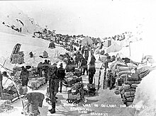 Peak of Chilkoot Pass in March–April 1898. Men wearing winter clothes with their supplies in the snow all of it surrounded by hill-sides.