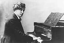 A man in his early 30s in a suit. He is sitting at a piano