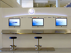 3 Apple computers, 2 with stools, installed on a white coloured stand by Oneworld