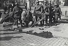 Seven soldiers of the German Army are stationed at a street corner in Helsinki after the surrender of the Red Guard headquarters Smolna. One of them is on his knee while two are relaxing against a railing or on a chair. MG 08, a heavy machine gun rests in front of them.