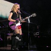 Image shows a blond woman in shorts, playing a purple electric guitar. She is shown wearing long, black socks on her legs.