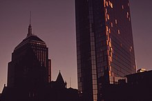 Two dark buildings rise into the early evening sky. The tower on the right, the Hancock Tower, has plywood on its side.