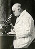 Elderly bespectacled gentleman sitting at a table and looking in a microscope