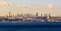 Skyline of a portion of the city, including several skyscrapers interspersed among low- and mid-rises, several historic buildings, parks and hilly terrain, and the waterway known as Bosphorus