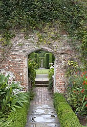 a view of an arch in a pink brick wall with a path leading to a statue