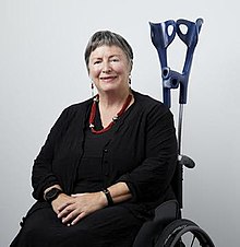 A white woman in black clothing sits in a wheelchair with crutches behind her