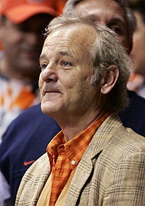 A three-quarters bust photograph of a 56-year-old white man with greying hair; he is wearing an orange collared shirt under a tan tartan blazer and intently looking to the left of the camera