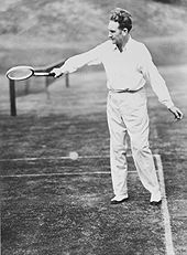 A man in white, with long pants and a shirt, swings a racket with his right hand on a grass court