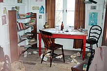 In the centre of a cluttered, whitewashed room stands a red table with two chairs, behind is a double window with simple brown curtains. Writing paraphernalia is strewn across the room