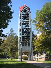 A three-dimensional vertical structure perhaps 70 feet (21 m) tall and 10 feet (3.0 m) wide by 10 feet (3.0 m) long is filled with tiers of large metal bells. The structure, standing in front of a large institutional building in a park-like setting, is supported by several metal legs. A sidewalk passes through the structure, which is open at the bottom to a height of perhaps 10 feet (3.0 m).