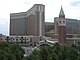 Distant ground-level view of a V-shaped building with a brick and tan facade and dark windows. Two shorter buildings with similar appearances are visible in the distance. A tall brick tower with a steep, green, pyramid-shaped roof is in the foreground.