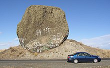 This photo shows an automobile passing in front of a rock which is essentially fully exposed. The rock has a rough, dark surface indicating it is weathered basalt and is roughly circular in exposed cross-section. The rock is immediately adjacent to a roadway – the road cut removed much of the earth from one side of it exposing it – from the excavation it is evident that the rock sits on a mound of glacial till. The rock is approximately 2 times the length of the car (i.e., ≈9 meters) in one direction and 5 times the height of the car in the other direction (i.e., ≈9 meters). Since the rock has not tipped onto the road and no structural support is provided, it must be approximately as deep as it is wide and high. Since the density of basalt is 3 grams per cubic centimeter, this puts the mass of the rock at about 400 to 500 metric tons (consistent with the references).