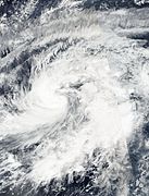 A tropical storm with clouds wrapping around the center; wind shear can be seen eroding the storm from the east