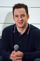 A mid shot of a man with curly brown hair, looking towards the camera and smiling. He is holding a microphone at a press event.